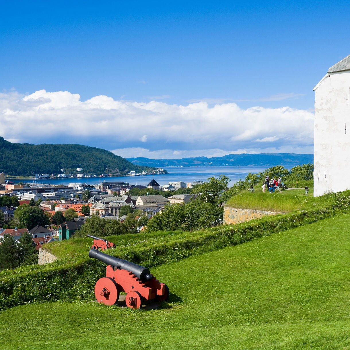 White tower of Kristiansten Fortress overlooking Trondheim city and fjord, with historic cannons on the grassy hillside under a bright blue sky.