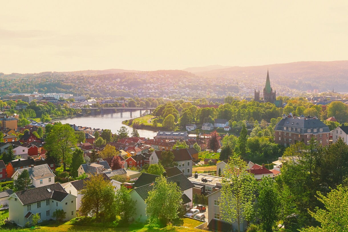 Panoramic view of Trondheim, Norway, with red and white houses, a river cutting through the city, and the spire of Nidaros Cathedral surrounded by trees at sunset.