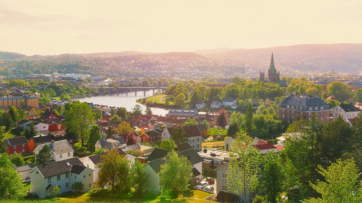 Panoramic view of Trondheim, Norway, with red and white houses, a river cutting through the city, and the spire of Nidaros Cathedral surrounded by trees at sunset.