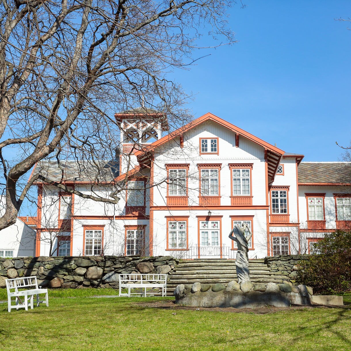 White wooden Ringve Museum with red trim in Trondheim, fronted by a stone wall, leafless trees and benches on a grassy lawn under a clear blue sky.