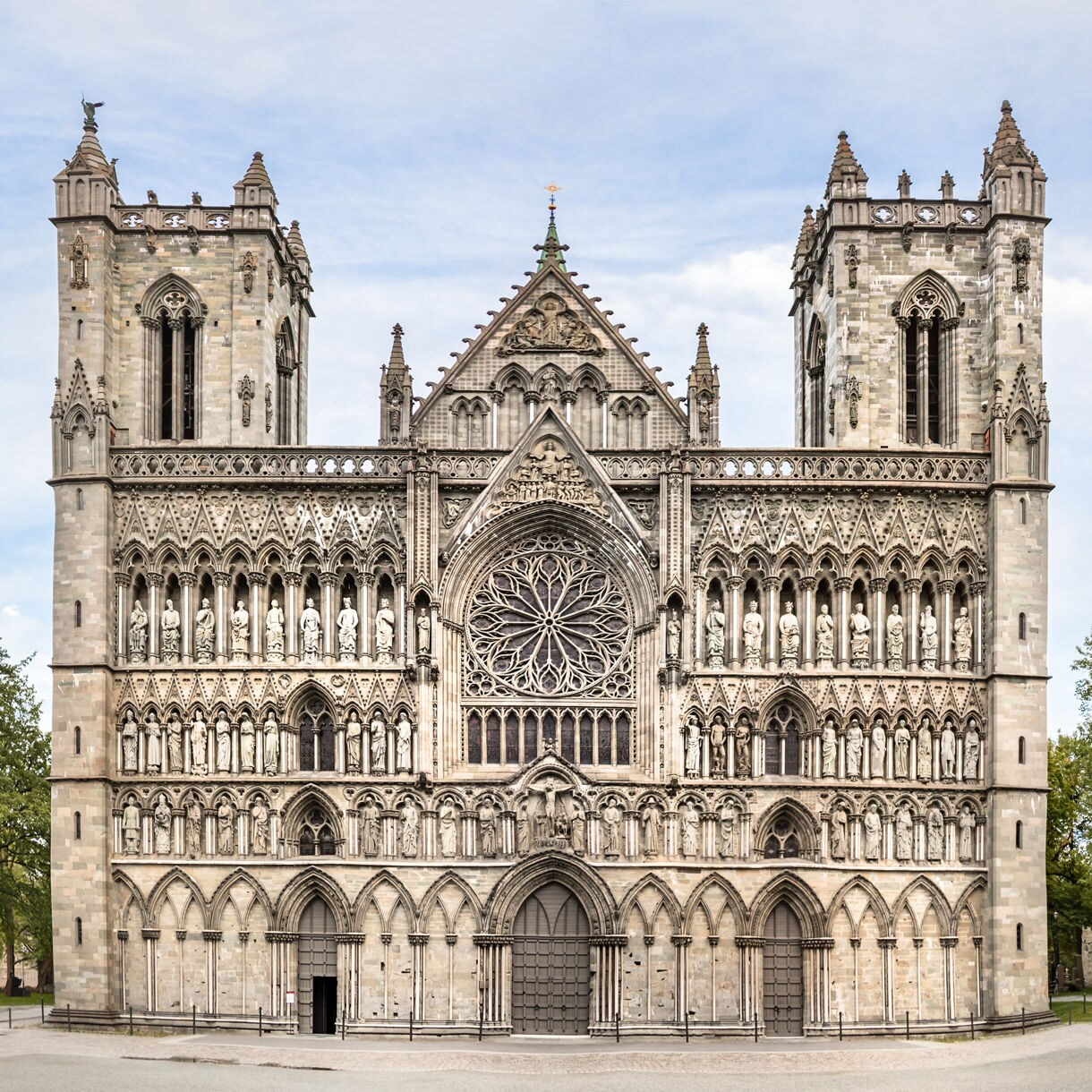 Front view of Nidaros Cathedral in Trondheim, featuring detailed Gothic carvings, twin towers and a large rose window surrounded by stone statues.