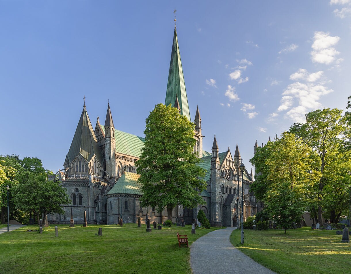 Exterior of Nidaros Cathedral in Trondheim with tall spires, a green copper roof and stone walls surrounded by trees and a grassy churchyard under a clear sky.