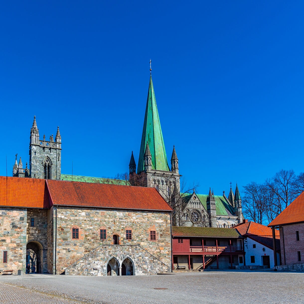 Stone Archbishop’s Palace with red-tiled roofs in Trondheim, Norway, with the spire of Nidaros Cathedral rising in the background under a vivid blue sky.