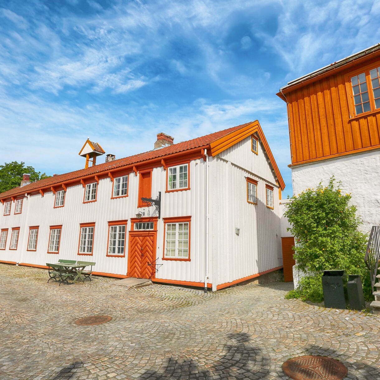 Courtyard view of the Ringve Museum in Trondheim with white and orange timber buildings, cobblestone paving and a vivid blue sky