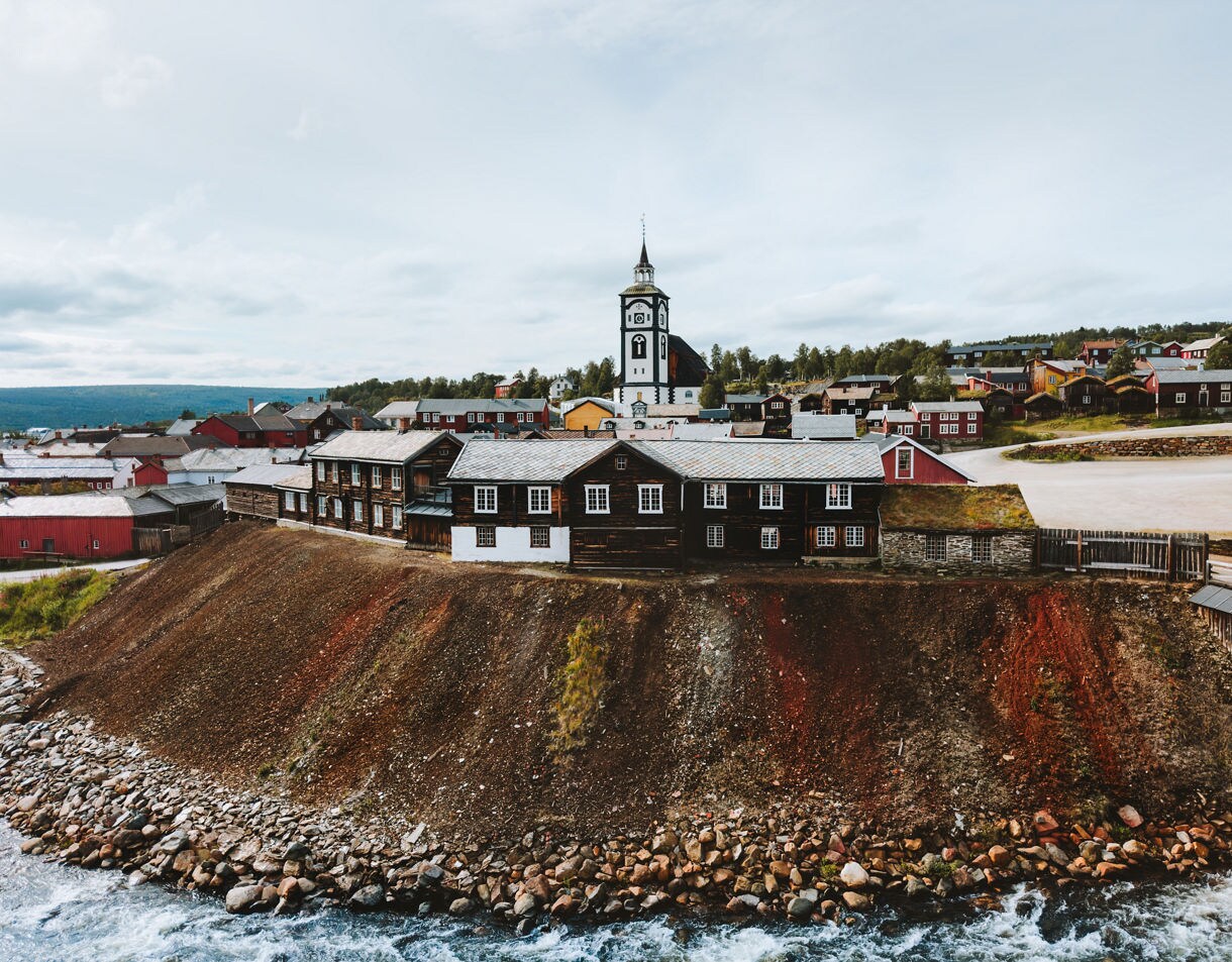 Traditional timber buildings and the white church tower of Røros, Norway, set on a hillside above a rocky riverbank under a cloudy sky.