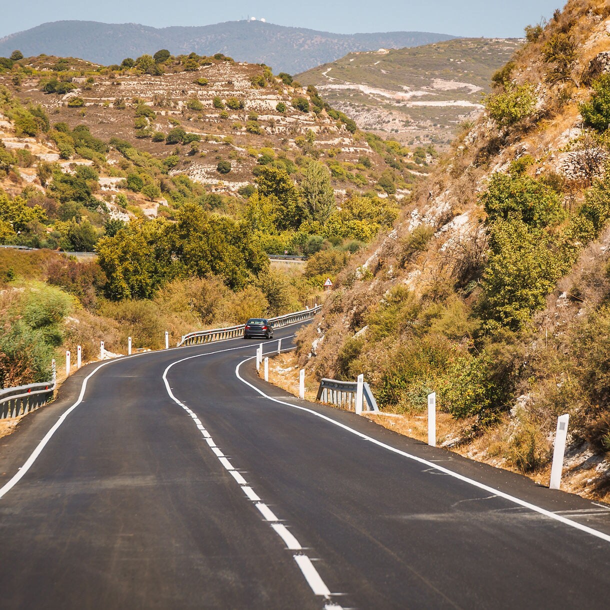 Curving mountain road in the Troodos region of Cyprus surrounded by rugged hills, rocky slopes and patches of green trees under a clear sky.