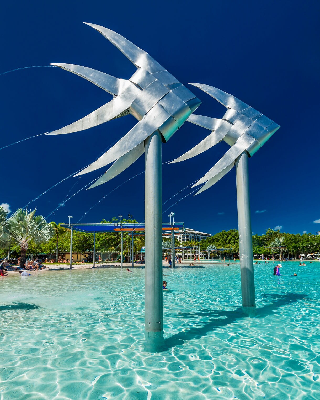 Shallow turquoise lagoon with metallic fish sculptures and people swimming under a deep blue sky at Cairns Esplanade.
