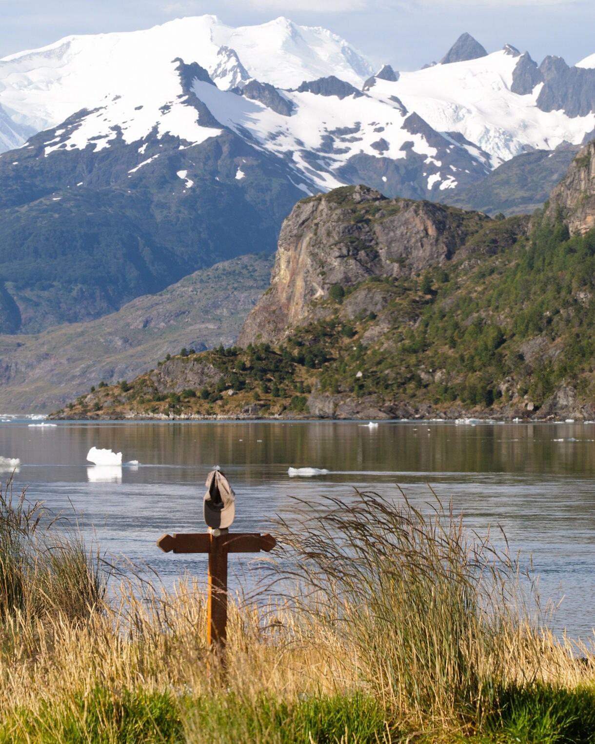 Grassy shoreline overlooking a still bay with floating ice, backed by rugged cliffs and snow-covered mountains under a soft, cloudy sky.