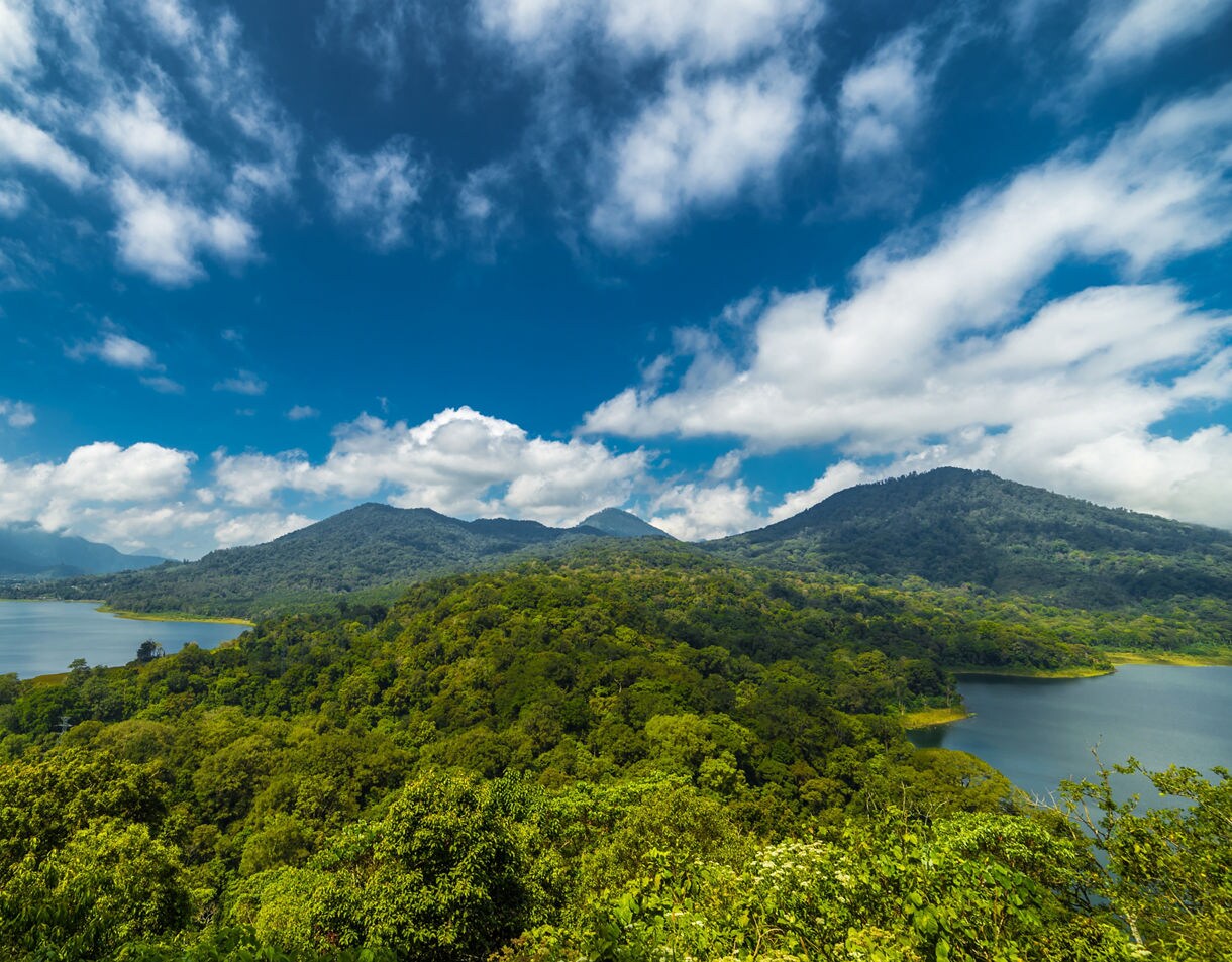 Panoramic view of two lakes surrounded by dense green forest, with rolling mountains rising in the background under bright blue sky and scattered white clouds.