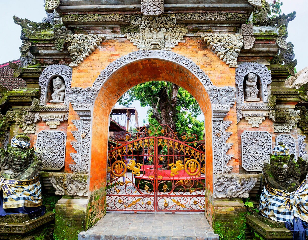 Intricate Balinese temple gate made of orange brick and detailed gray stone carvings, featuring statues wrapped in checkered cloth, a decorative red and gold iron gate and lush greenery visible through the arched entrance.