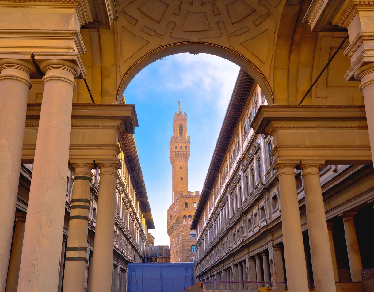 A view through the Uffizi Gallery’s columned courtyard toward the tall brick tower of Palazzo Vecchio, with warm stone architecture and a blue sky above.