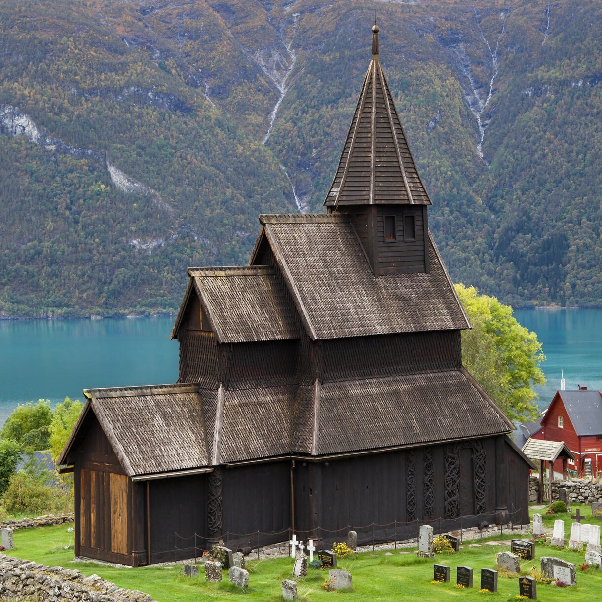 Urnes Stave Church, a dark wooden medieval church with carved details and steep roofs, stands on a hillside graveyard above a turquoise fjord with mountains in the background.