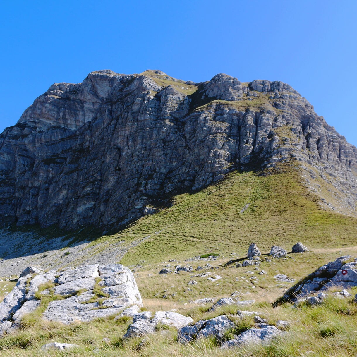 Uvita Greda mountain in Montenegro with steep rocky cliffs and grassy lower slopes, under a clear blue sky.