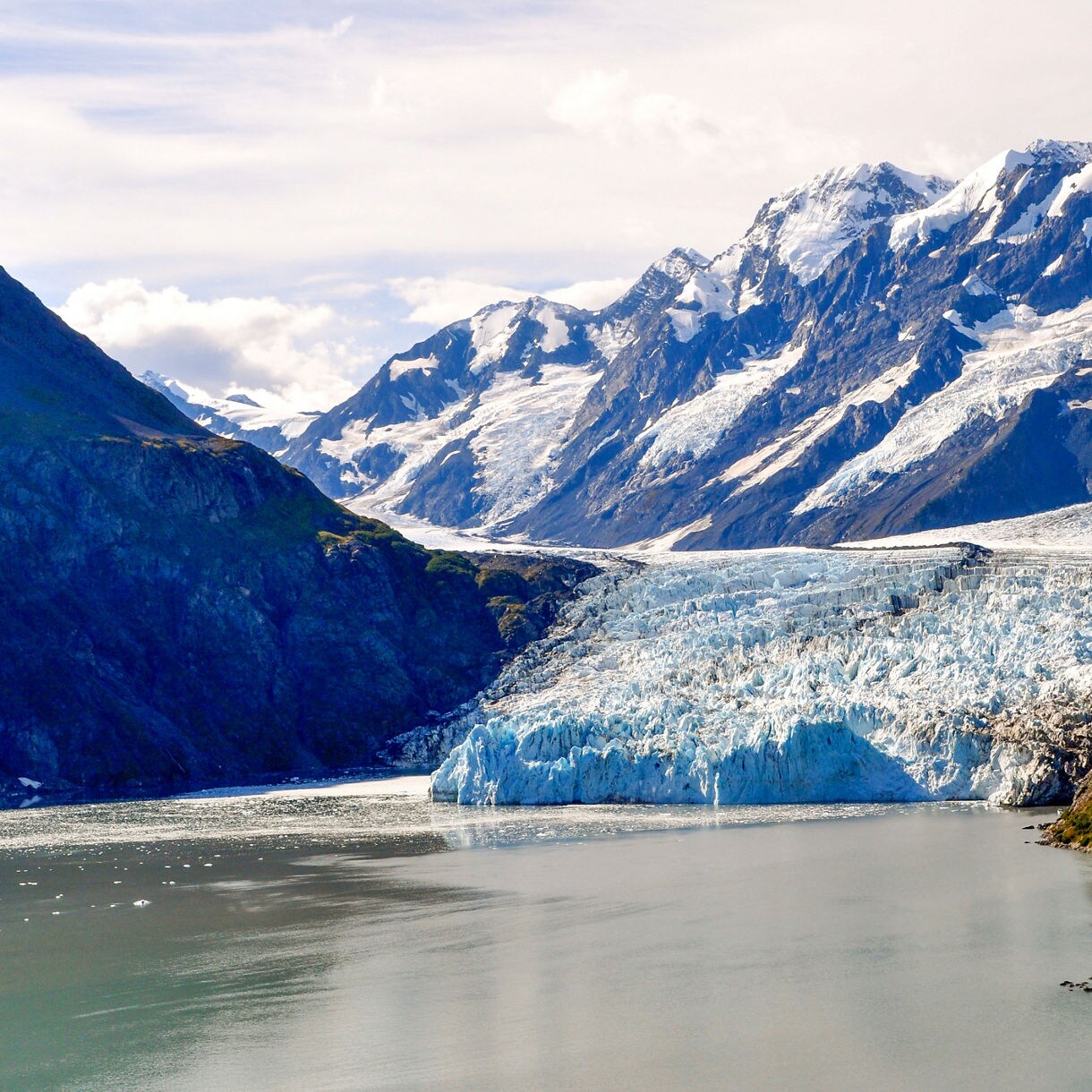 A massive glacier flows down between rugged mountain peaks, ending at the edge of a calm lake. Snow-capped summits rise in the background, contrasting with the icy blue of the glacier and the green slopes. The scene is bathed in natural daylight, creating a crisp and serene atmosphere. No visible text or numbers are present in the image.