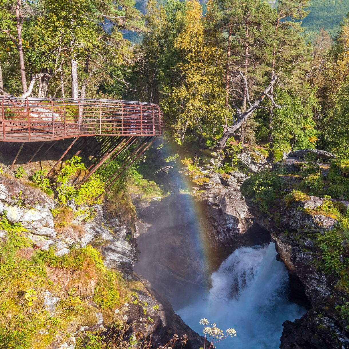 Waterfall cascading through rocky cliffs in Valldalen Valley with a rainbow visible in the mist and a viewing platform above.