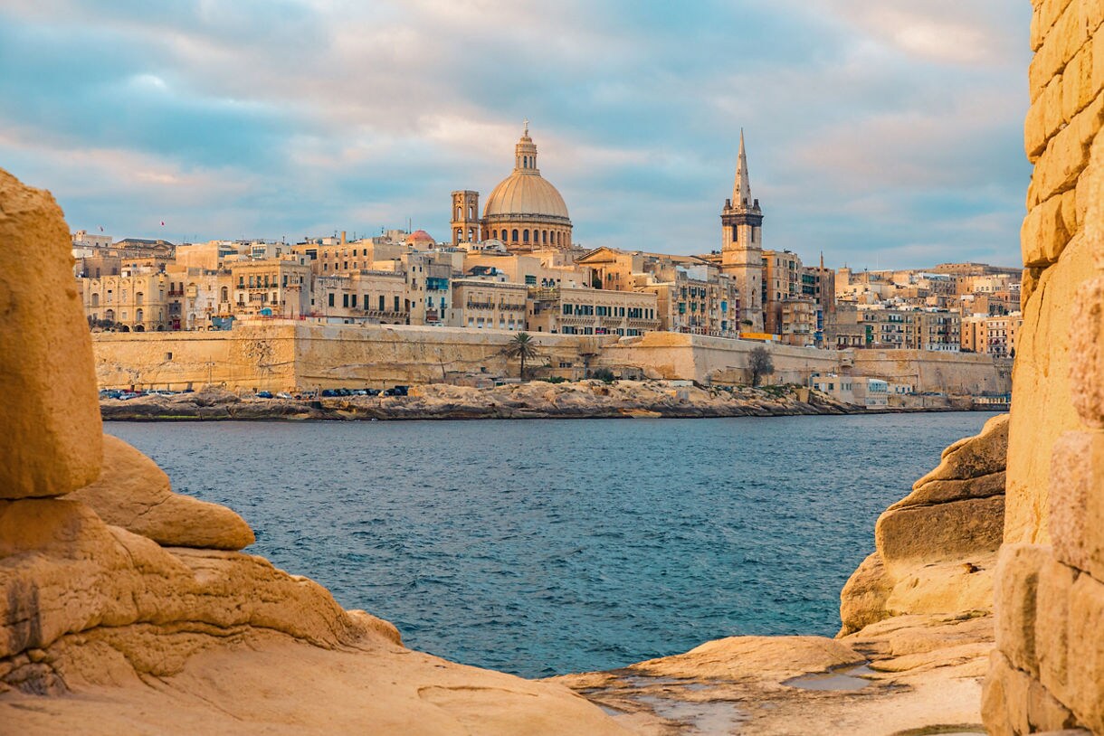 View of Valletta, Malta, with historic limestone buildings, a large dome and church spire overlooking the harbor at sunset.