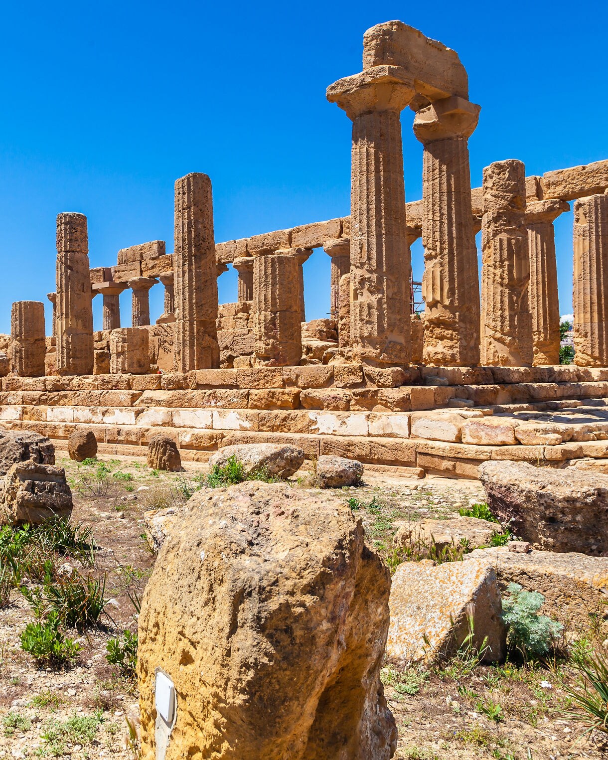 Ruins of a Greek temple in the Valley of the Temples in Agrigento, with tall weathered stone columns set on a rocky hillside under a bright blue sky.