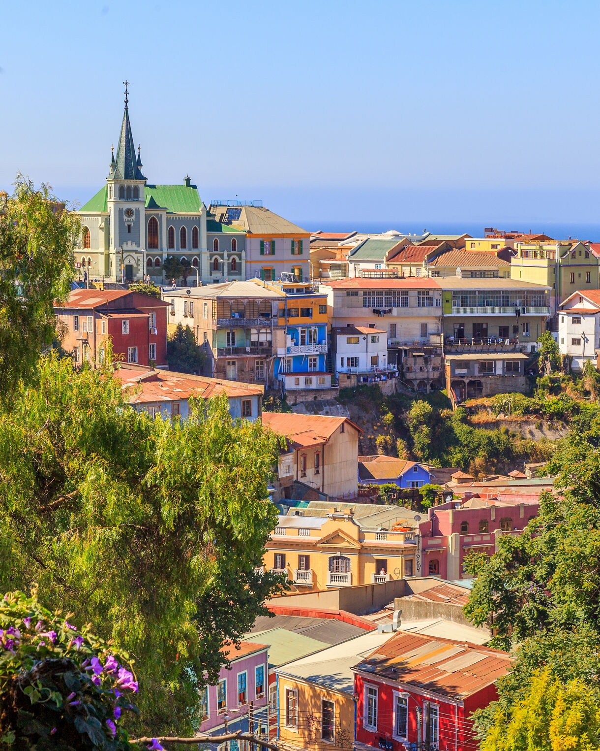 Vibrant houses stacked on Valparaíso’s steep hills with a church tower, green trees and a cargo ship on the blue ocean below.