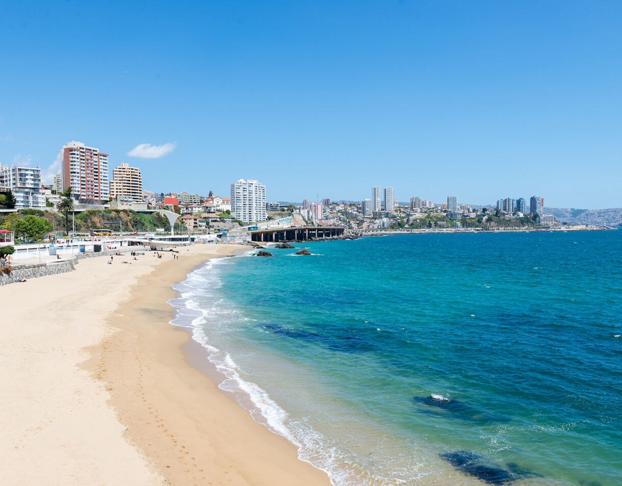 Wide sandy beach in Valparaíso Bay with turquoise water and coastal city buildings rising along the shoreline.