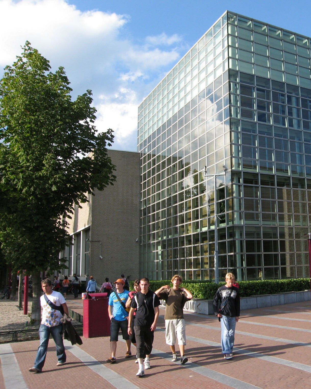 Exterior view of the Van Gogh Museum in Amsterdam featuring a modern glass façade, a brick annex and visitors walking across a wide plaza under a bright blue sky.