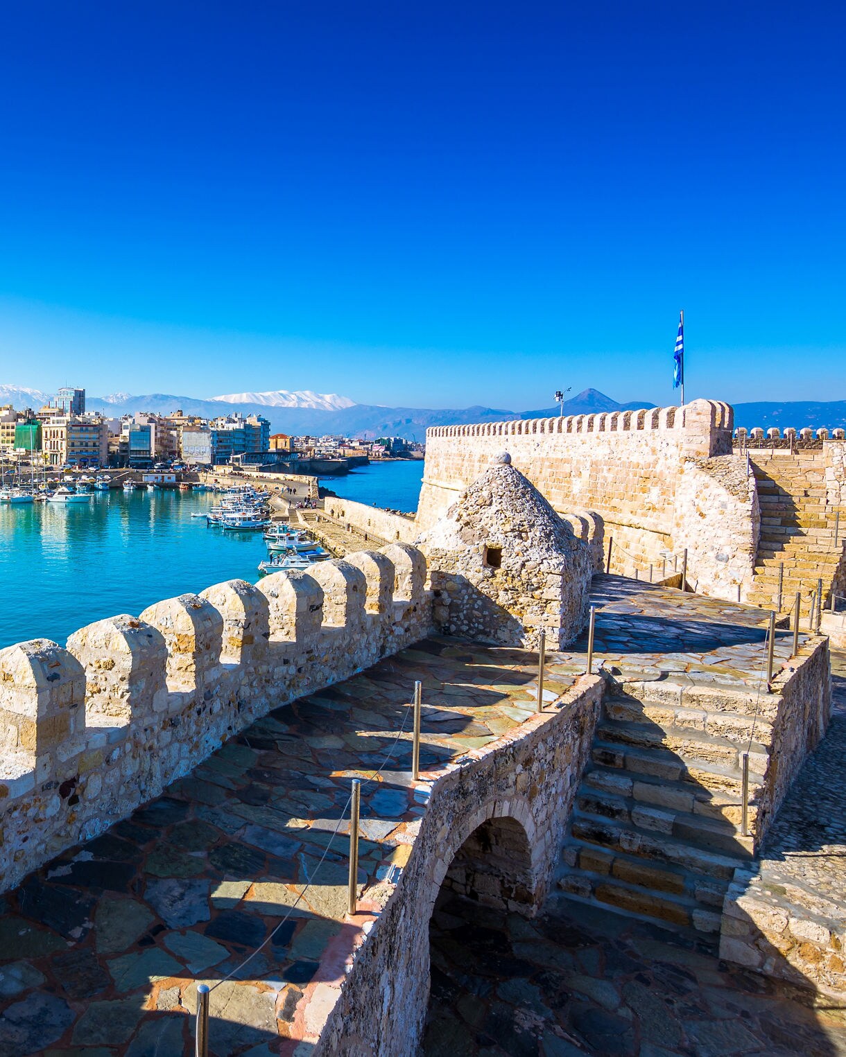 View of Heraklion’s Venetian harbor fortress overlooking a marina filled with boats, with the city skyline and distant mountains under a clear blue sky.