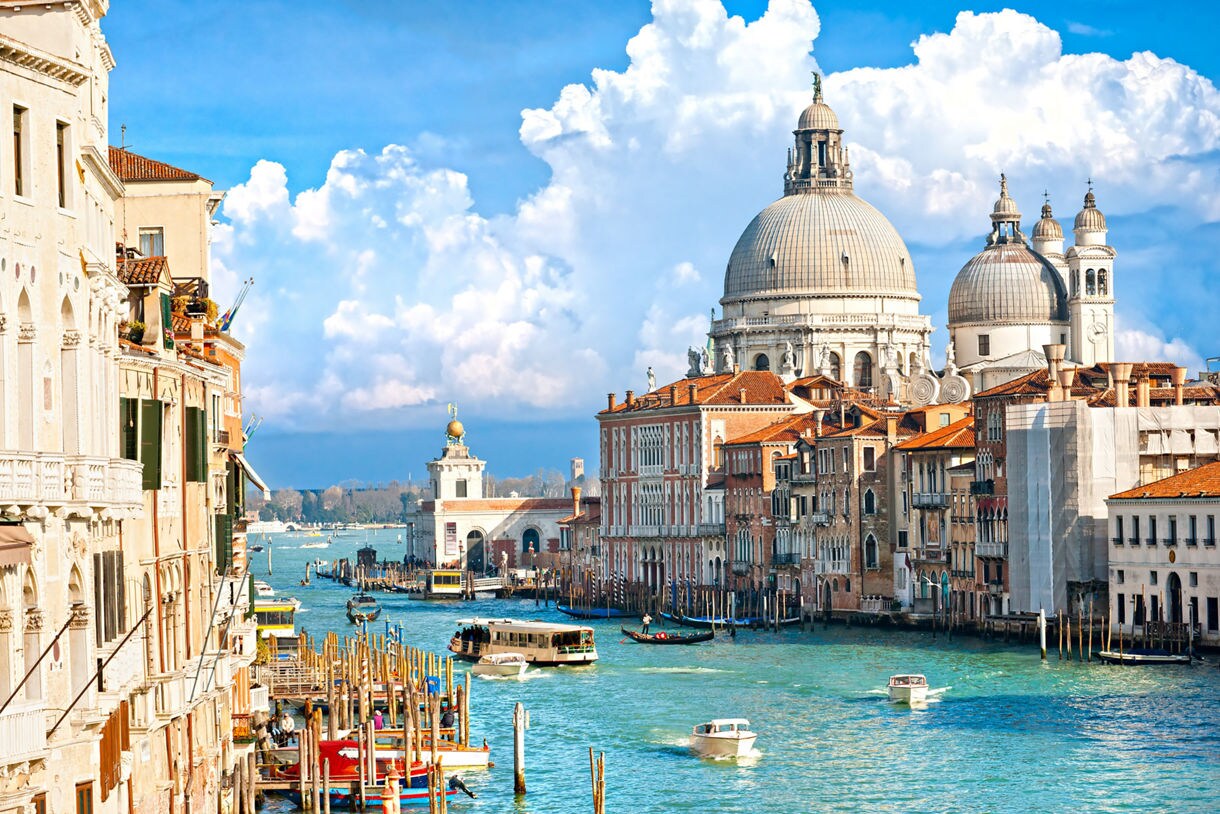 A stunning view of the Grand Canal in Venice Italy featuring historic buildings on either side, including the massive domes of Santa Maria della Salute church on the right, under a bright blue sky with large white clouds. Small boats and water taxis navigate the bright turquoise water.