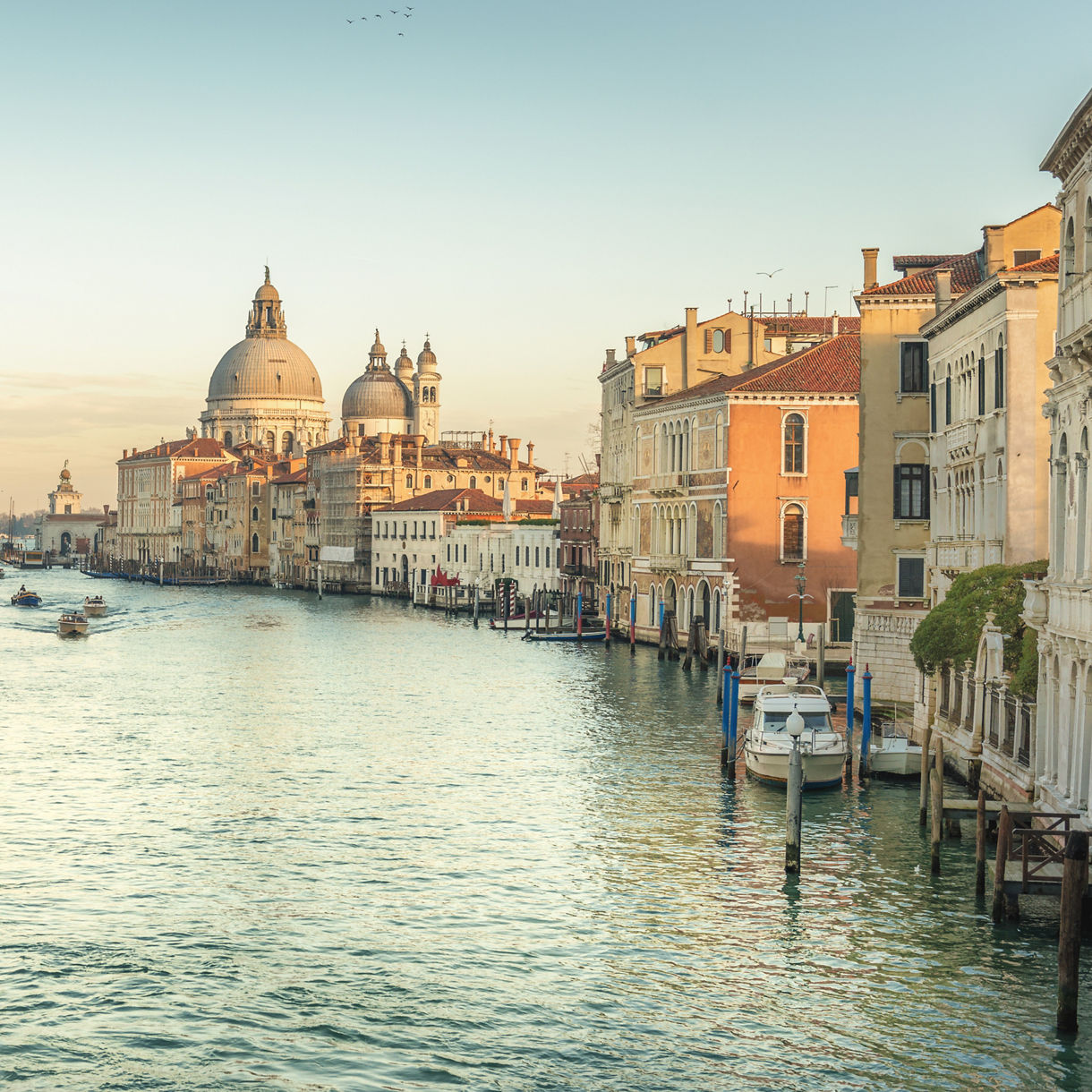 A panoramic view of Venice's Grand Canal at sunrise, with a lone gondola silhouetted on the calm water, historic palazzos lining both sides and the domed Santa Maria della Salute basilica glowing in the distance.
