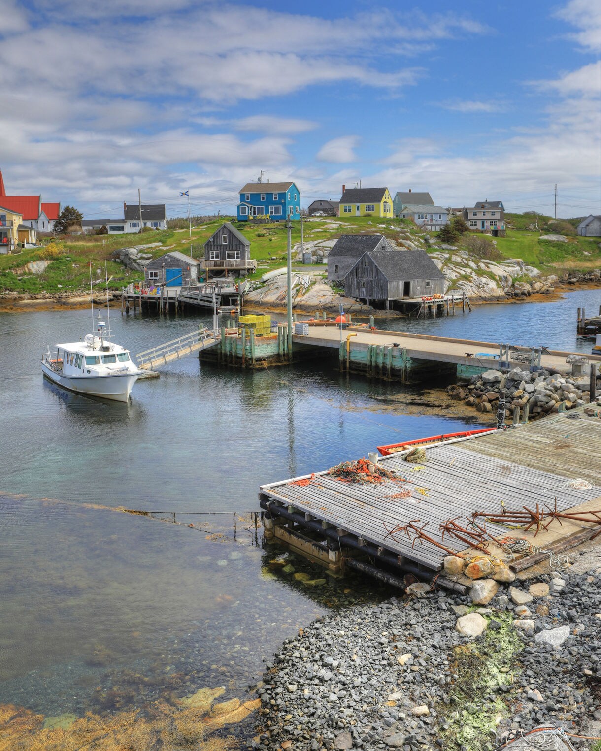 Small fishing harbor at Peggy’s Cove with a white boat near wooden docks, colorful hillside homes and weathered shacks set among rocky terrain under a partly cloudy sky.