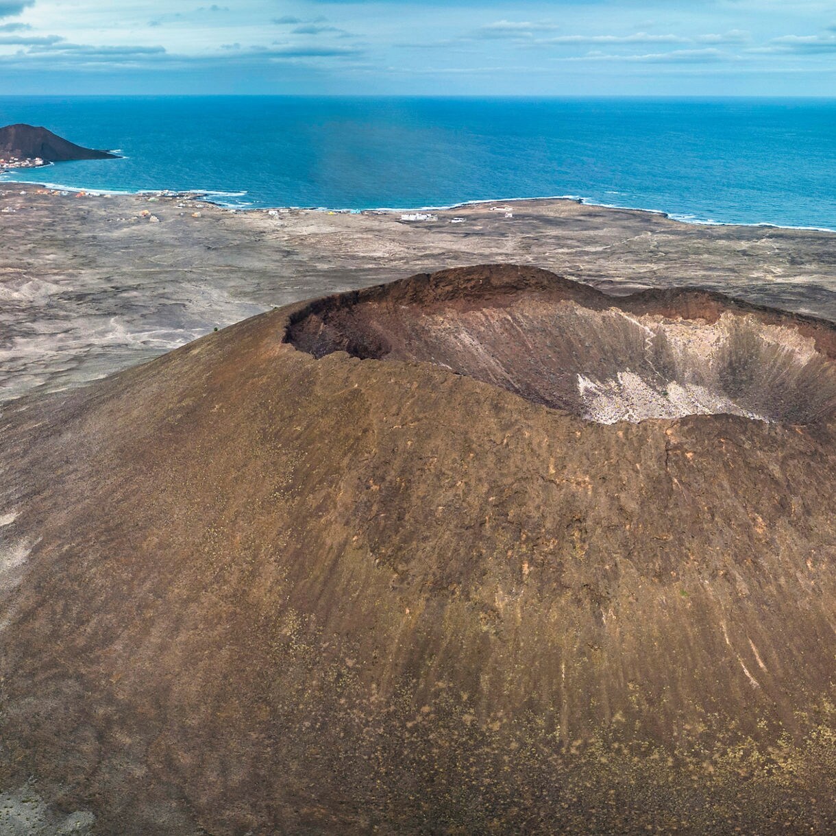 Aerial view of the Viana volcano on Cape Verde with a wide, steep-walled crater surrounded by barren volcanic terrain, distant hills and the deep blue ocean under a lightly clouded sky.