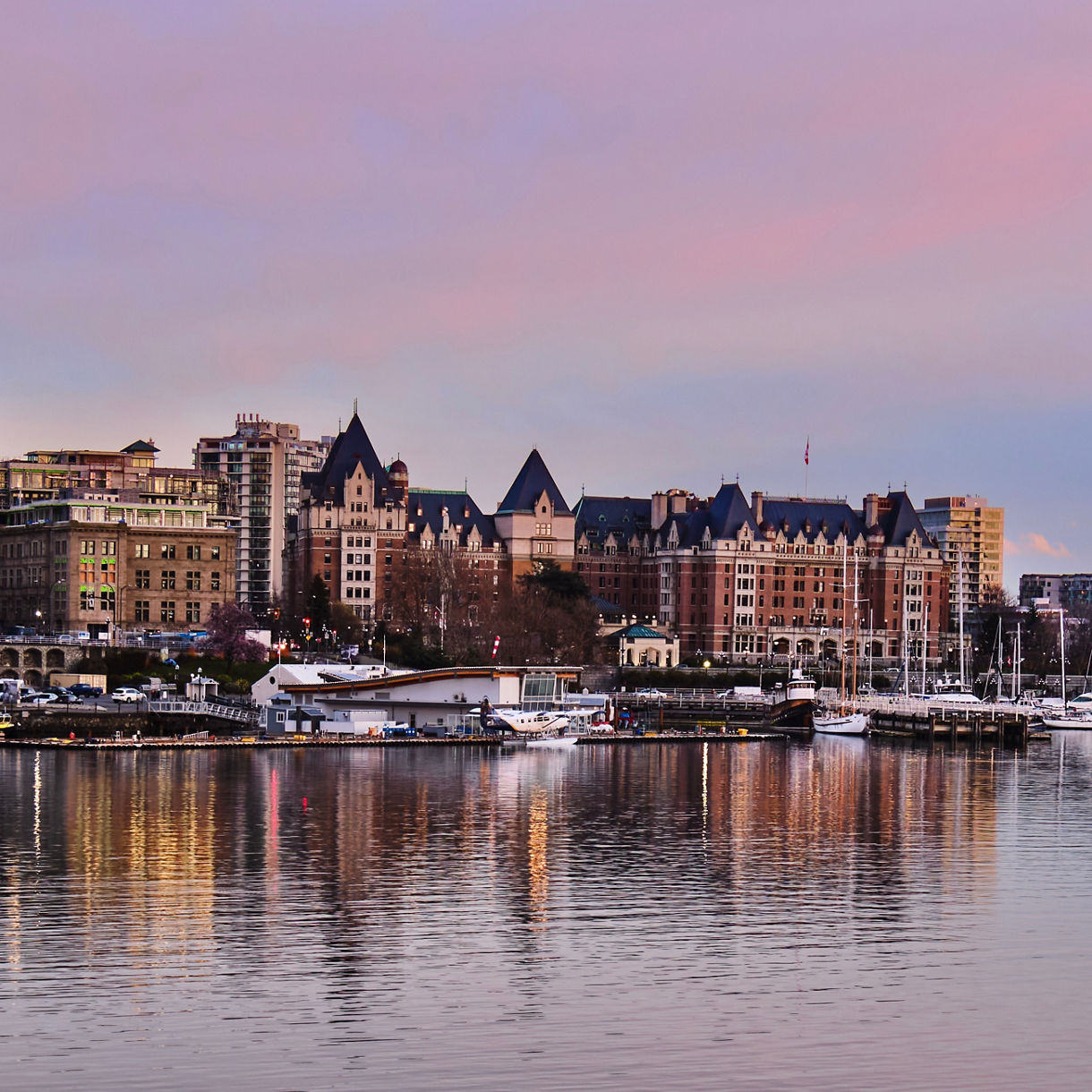 Panoramic view of Victoria's Inner Harbour skyline at dusk with a pink and purple sky, historic buildings and boats reflected in calm water.