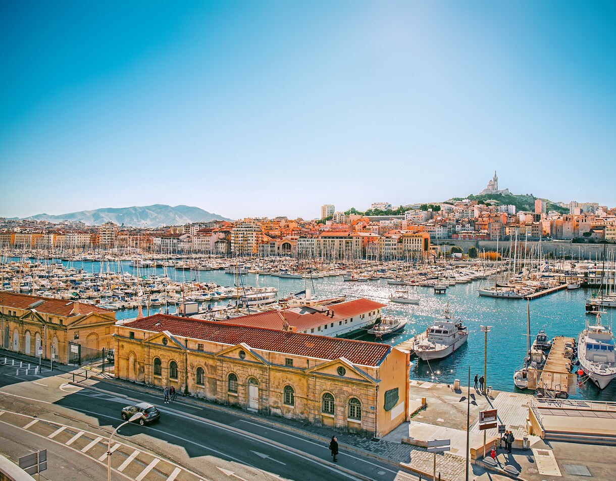 Panoramic view of Marseille’s Vieux Port featuring rows of sailboats, historic yellow buildings along the waterfront and the hilltop Notre-Dame de la Garde basilica under a clear sky.