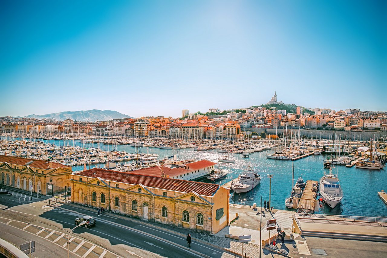Wide view of Marseille’s Vieux-Port with rows of boats and yachts docked in the marina, surrounded by terracotta-roofed buildings and overlooked by the hilltop basilica of Notre-Dame de la Garde.