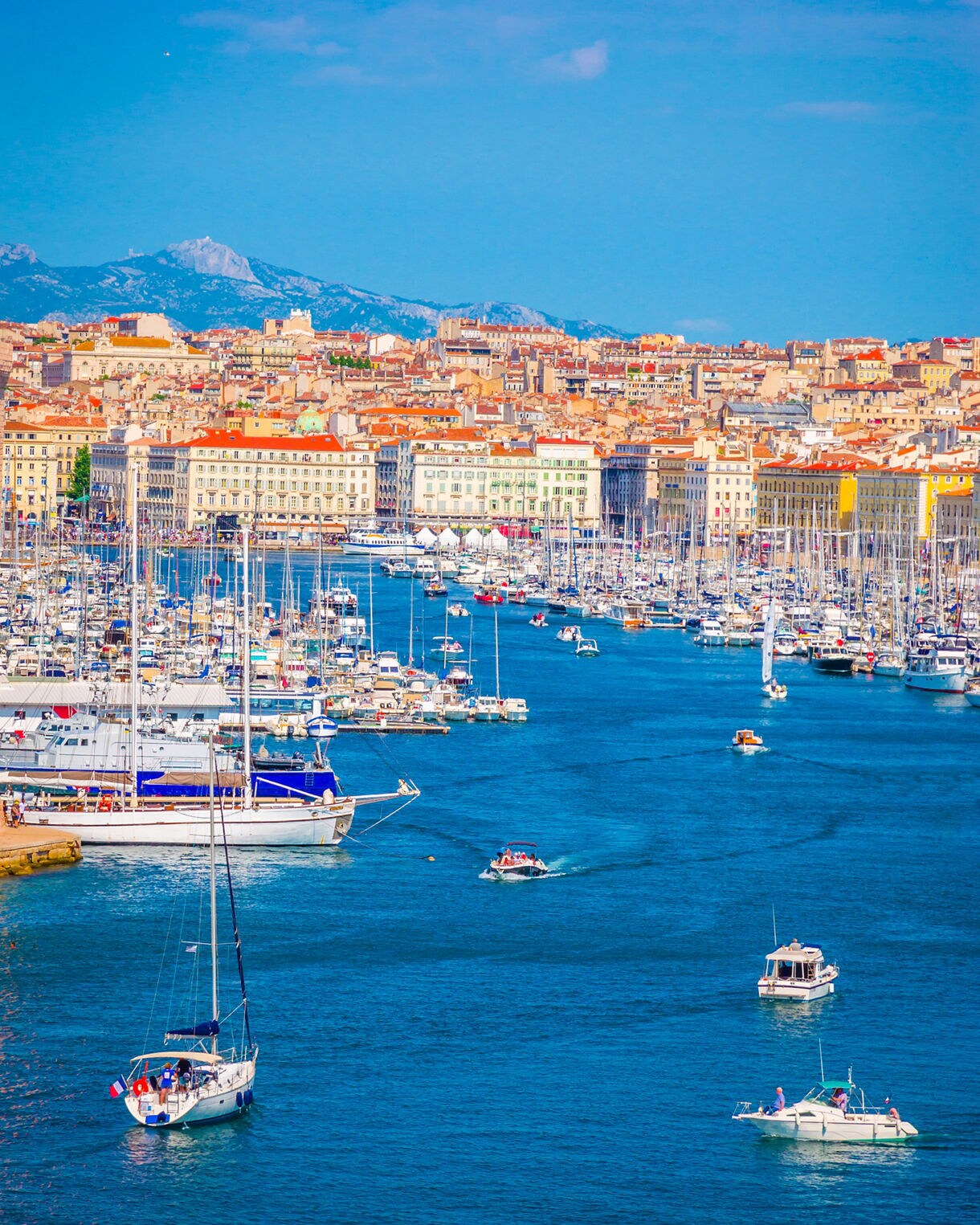 Colorful view of Marseille’s Old Port filled with sailboats and yachts, framed by historic buildings with terracotta roofs and distant mountains.