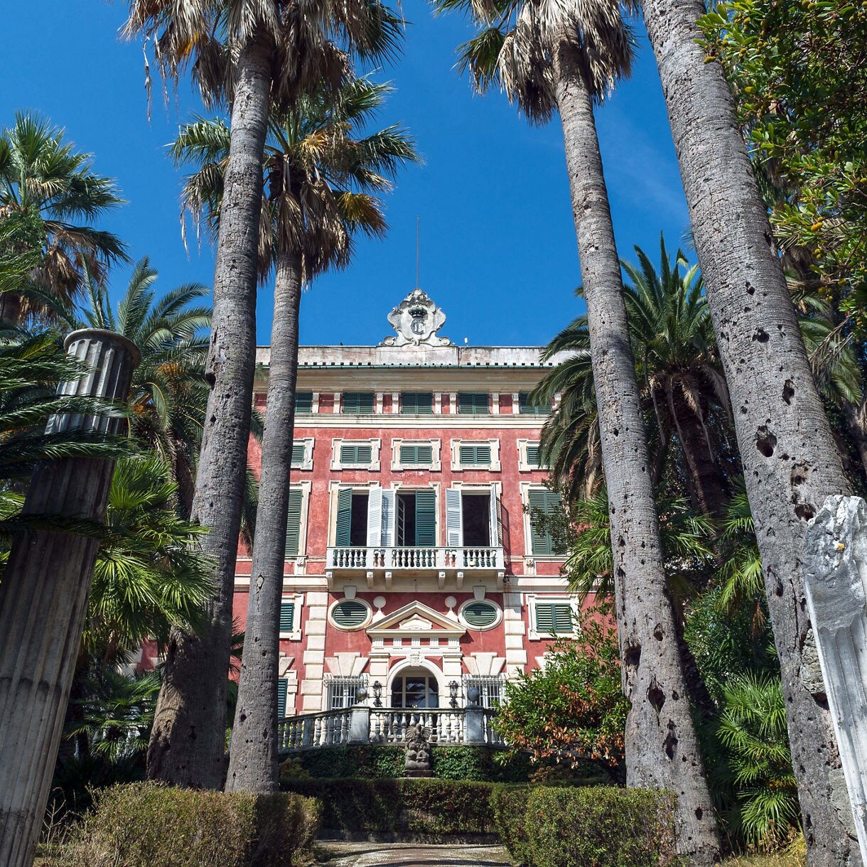 A historic pink villa framed by tall palm trees and dense greenery, with a small staircase and garden path leading to its entrance.
