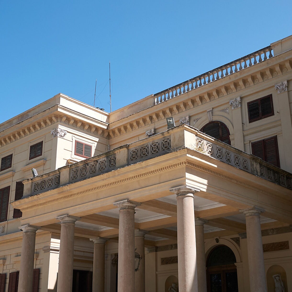 Historic villa with cream-colored walls, tall columns and decorative balconies under a clear blue sky.