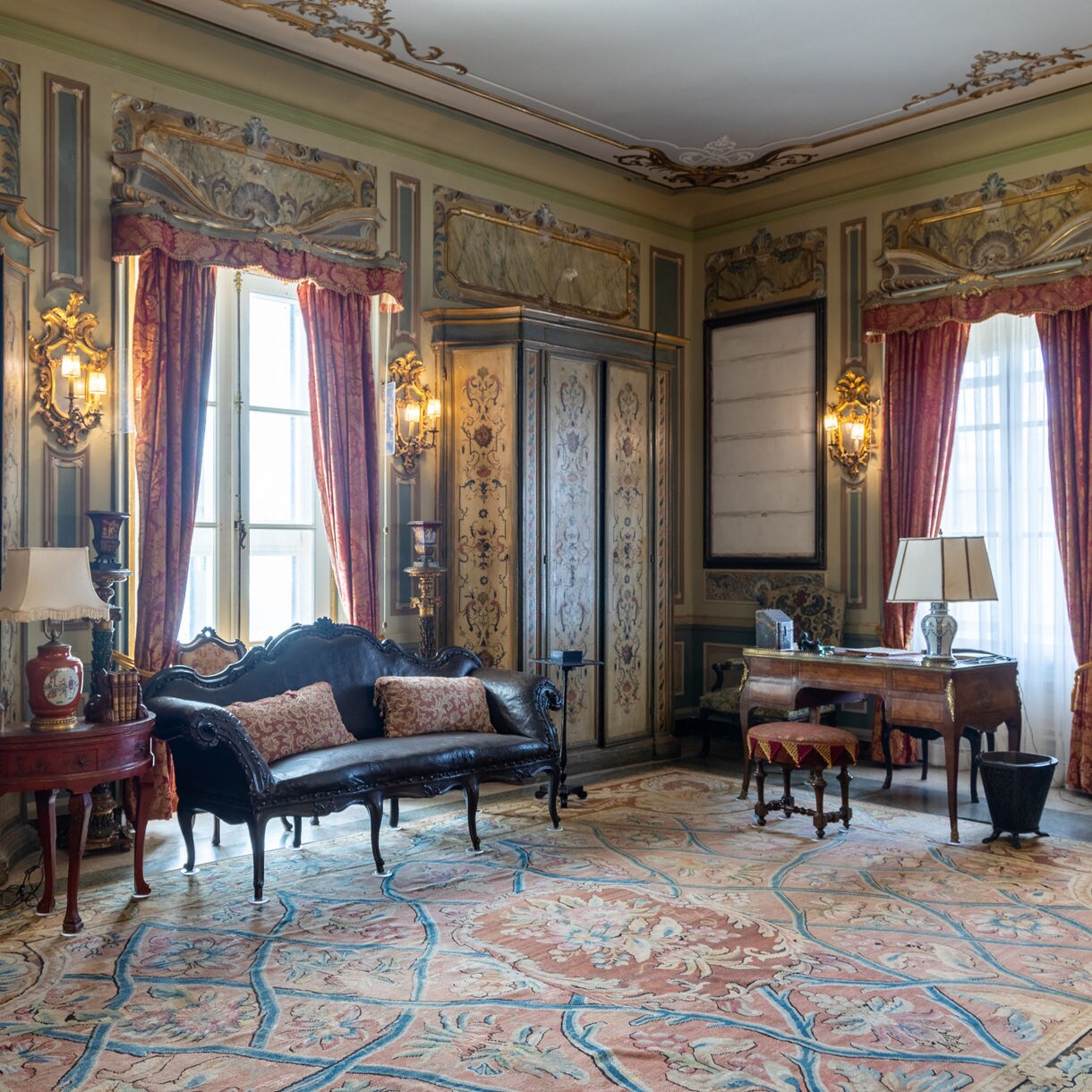 Elegant room inside Villa Vizcaya Museum in Miami, decorated with antique furniture, ornate gold-trimmed walls, red curtains and a large patterned rug.