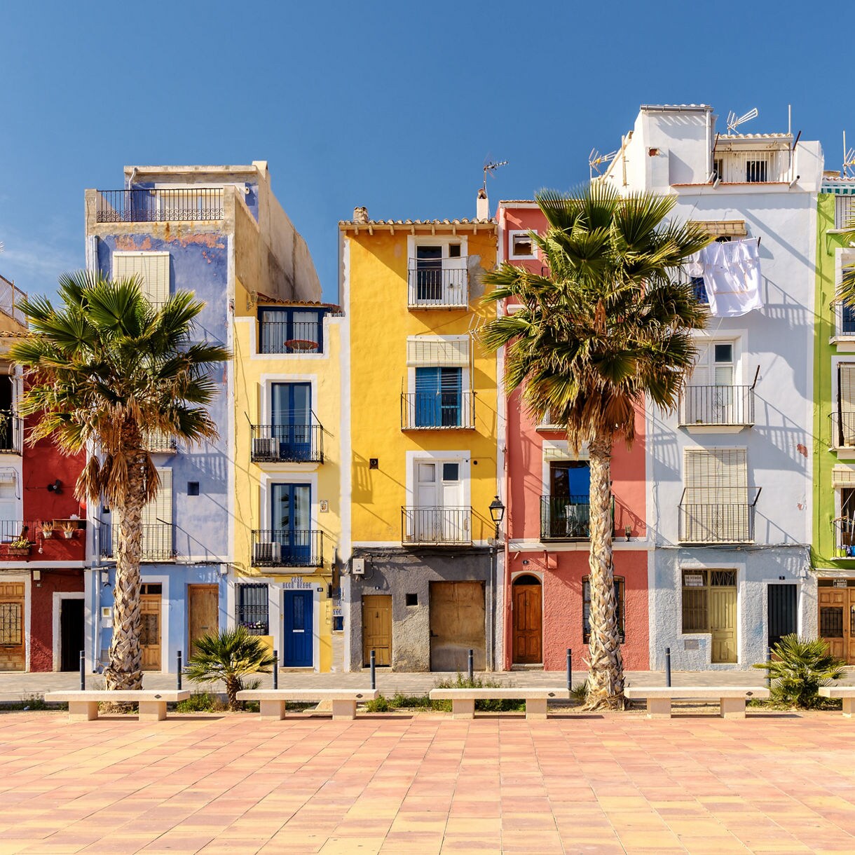 Row of tall pastel houses painted in vibrant shades of yellow, blue, pink and green framed by palm trees along a sunny coastal promenade in Villajoyosa, Spain.