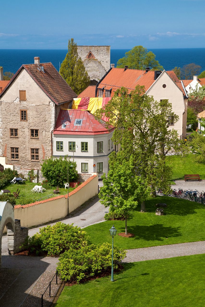 Historic stone houses and red rooftops in Visby with gardens, cobblestone paths and the Baltic Sea in the background.