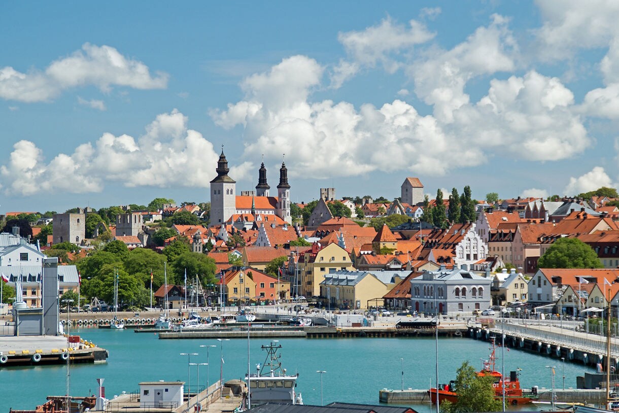 Panoramic view of Visby, Gotland in Sweden, with historic church towers, medieval stone ruins, red-tiled rooftops and a turquoise harbor in the foreground.