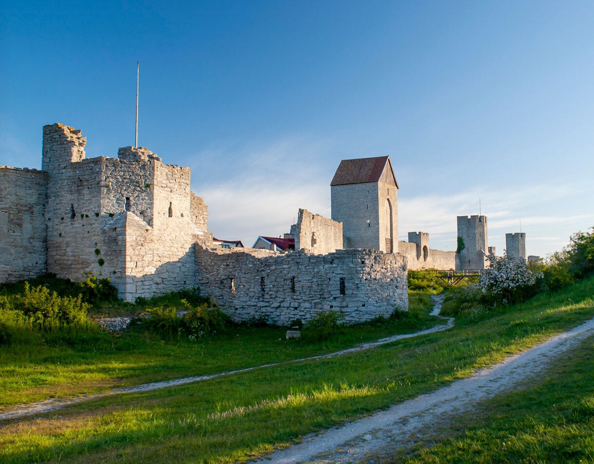 Stone ruins and towers of the medieval city wall in Visby, Gotland, Sweden, set against green grass and a clear blue evening sky.