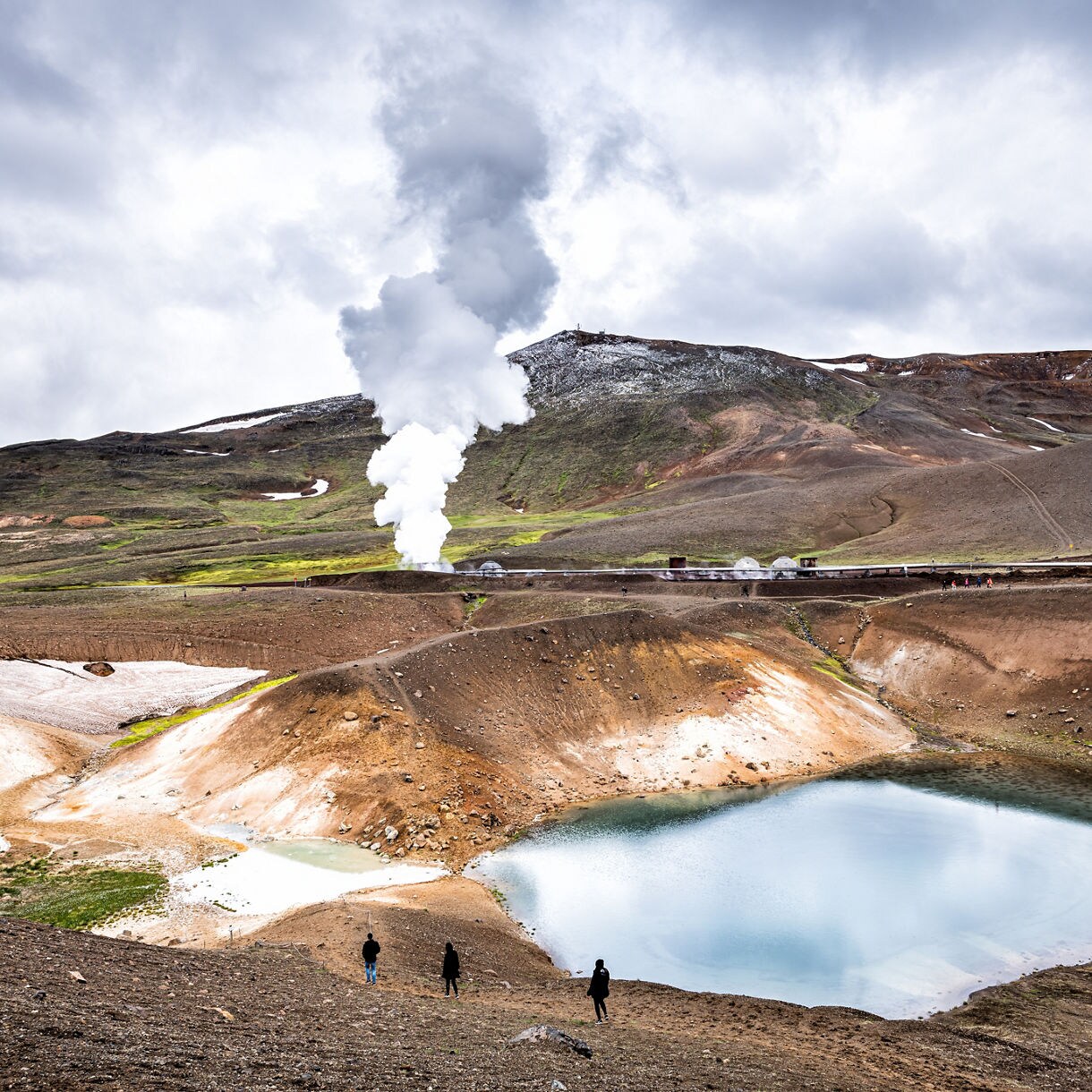 Víti Crater in the Krafla volcanic caldera, Iceland, featuring a blue crater lake surrounded by barren slopes with geothermal steam rising in the background under cloudy skies.