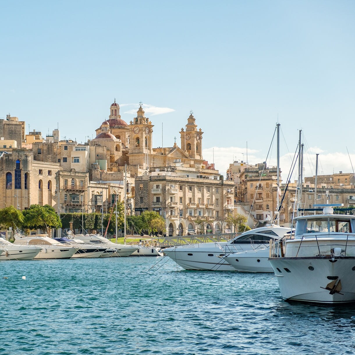 View of Vittoriosa harbor in Malta, with luxury boats docked along turquoise waters and historic limestone buildings topped by church domes in the background.