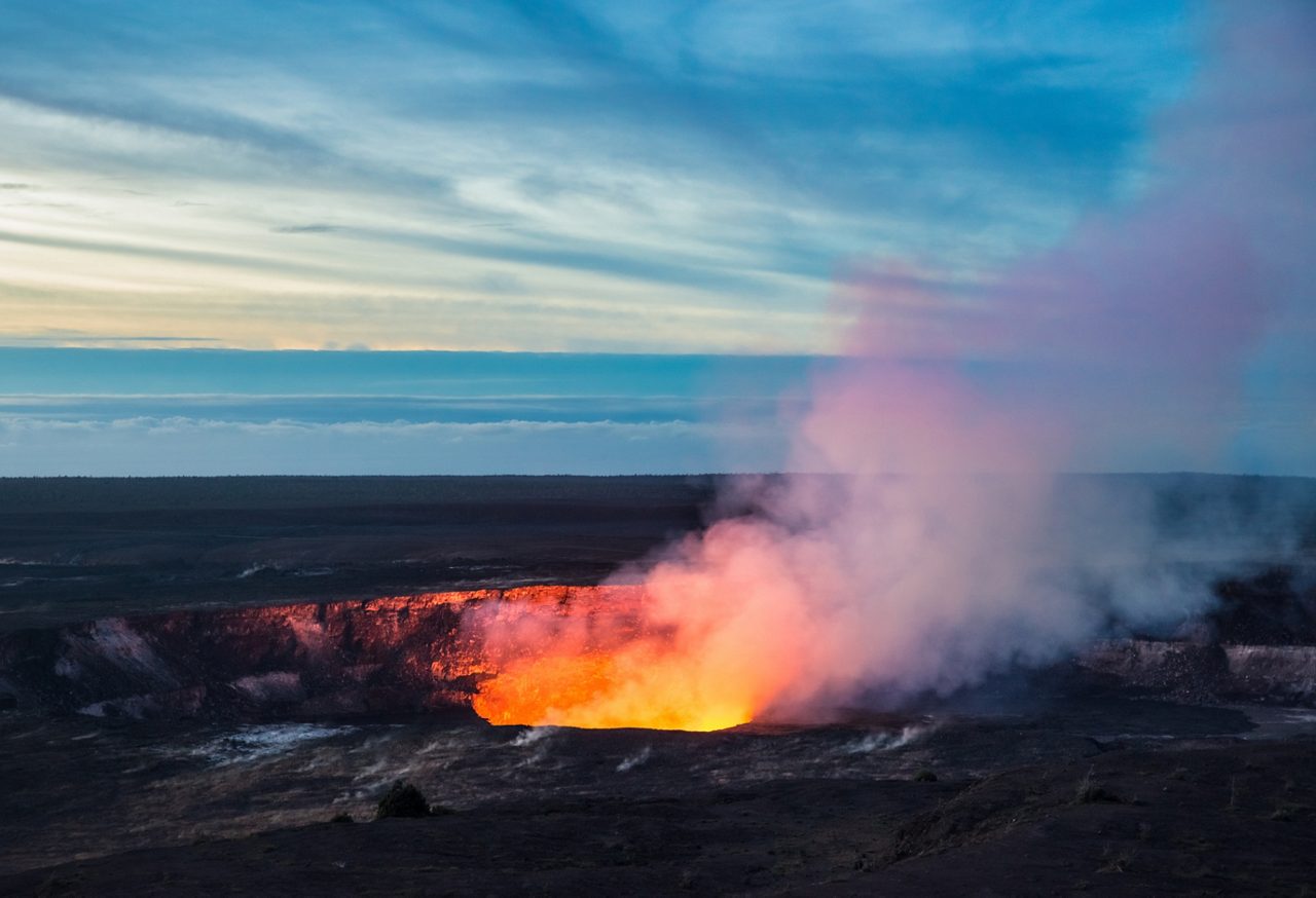 Bright orange lava and rising steam from Halemaʻumaʻu Crater inside Hawaiʻi Volcanoes National Park at dusk.