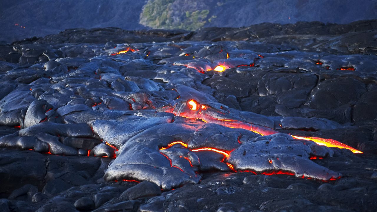 Flowing red lava weaving through hardened black rock with a glowing sunset sky above, where a rainbow arcs across the horizon.