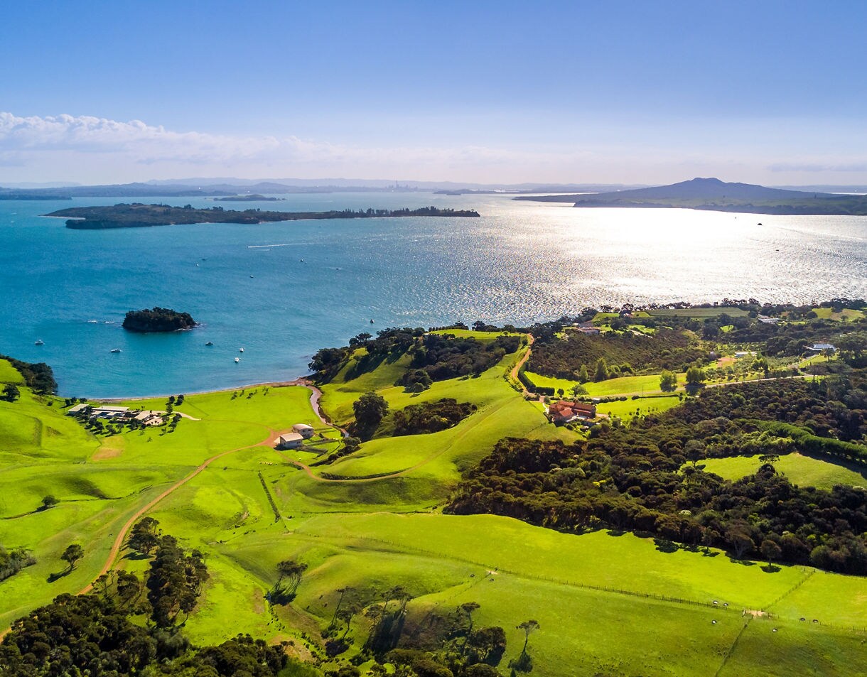 Aerial view of Waiheke Island with lush green fields, scattered homes and bright turquoise water under clear skies.