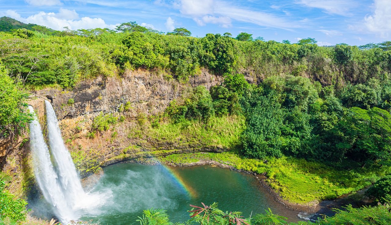 Wailua Falls in Kauai plunging into a green pool surrounded by dense tropical vegetation, with a rainbow forming in the mist.