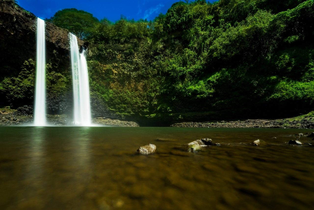 Twin waterfalls at Wailua Falls cascading into a tranquil pool, framed by lush green vegetation and moss-covered cliffs under a bright blue sky.