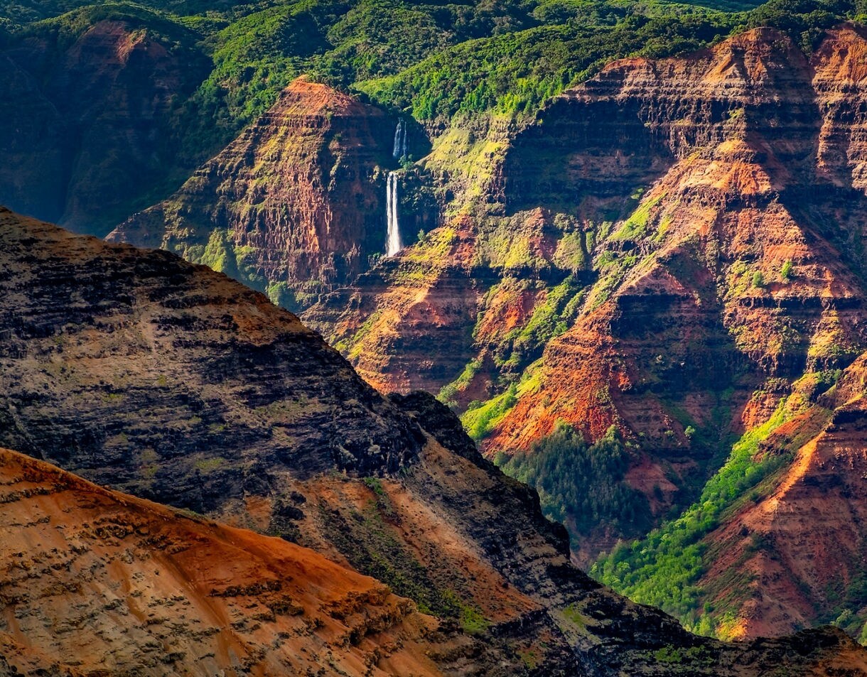 Panoramic view of Waimea Canyon on Kauai, showing deep red rock walls, green valleys and the Pacific Ocean in the distance under a bright blue sky.