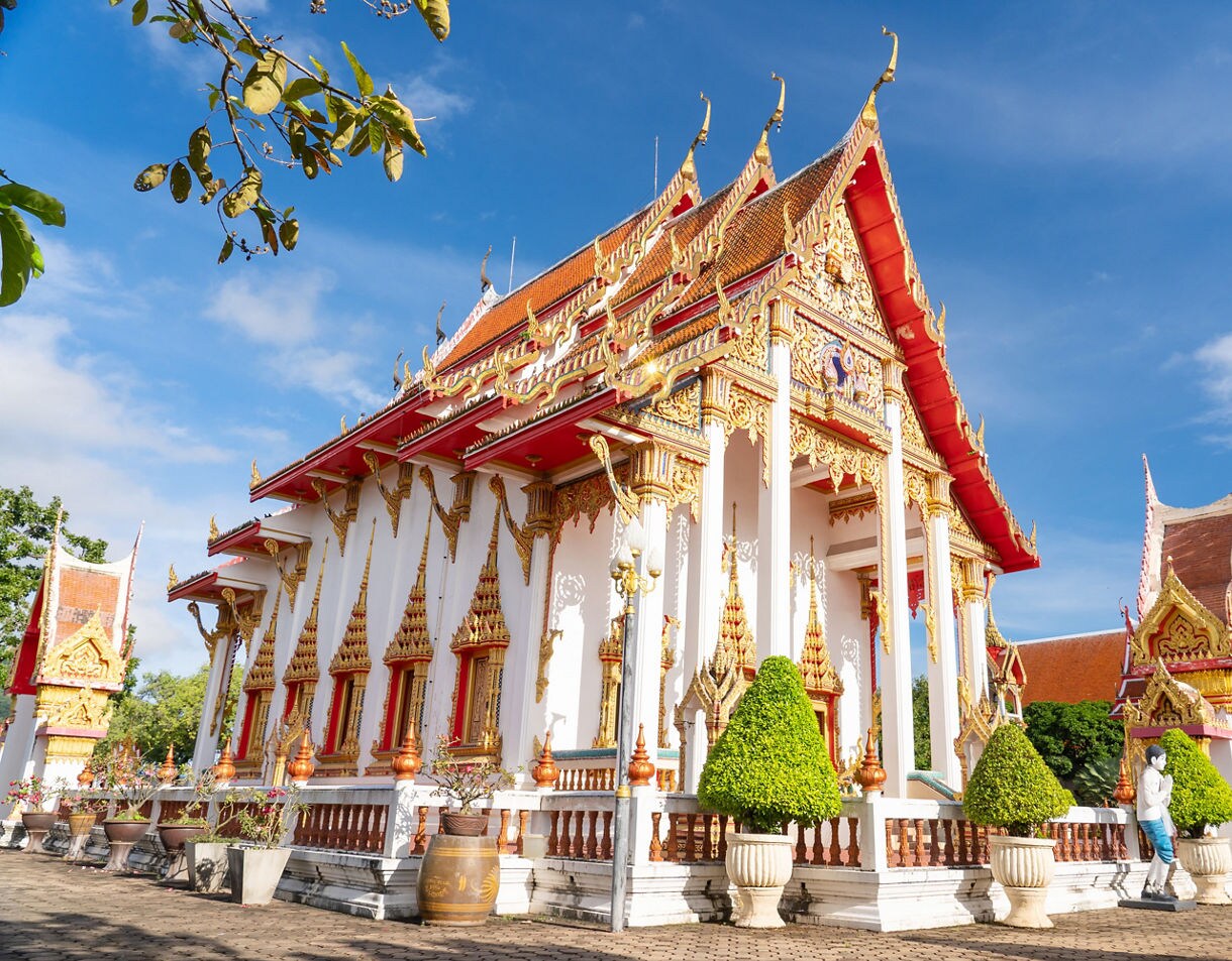 Ornate Thai temple at Wat Chalong with white walls, red and gold roofs and decorative spires shining under a clear blue sky.