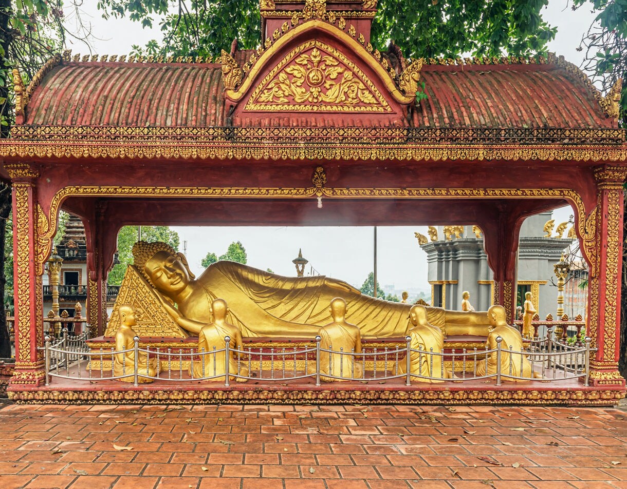 Golden reclining Buddha statue displayed under an ornate red and gold pavilion with small monk figures surrounding it.